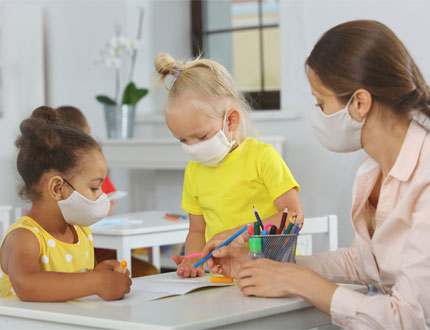 Early Head Start Young Girls With Teacher With Mask At Desk Photo